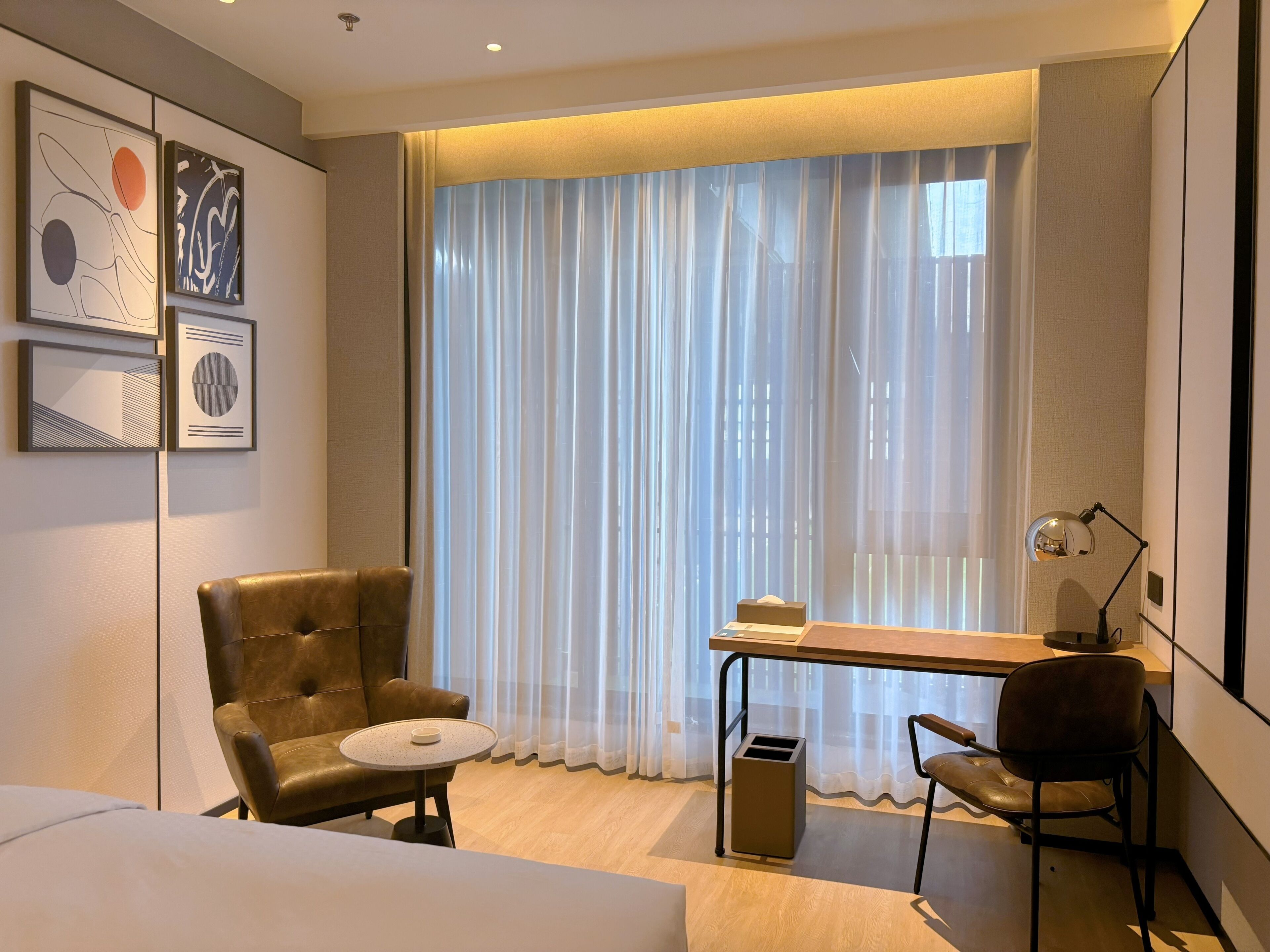 A modern hotel room with a large window, a leather armchair, a small round table, a desk with a lamp, and a trash bin.