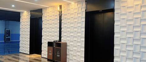 Un vestíbulo moderno con piso de mármol, una columna alta con lámpara y dos ascensores con un diseño de pared texturizado único.