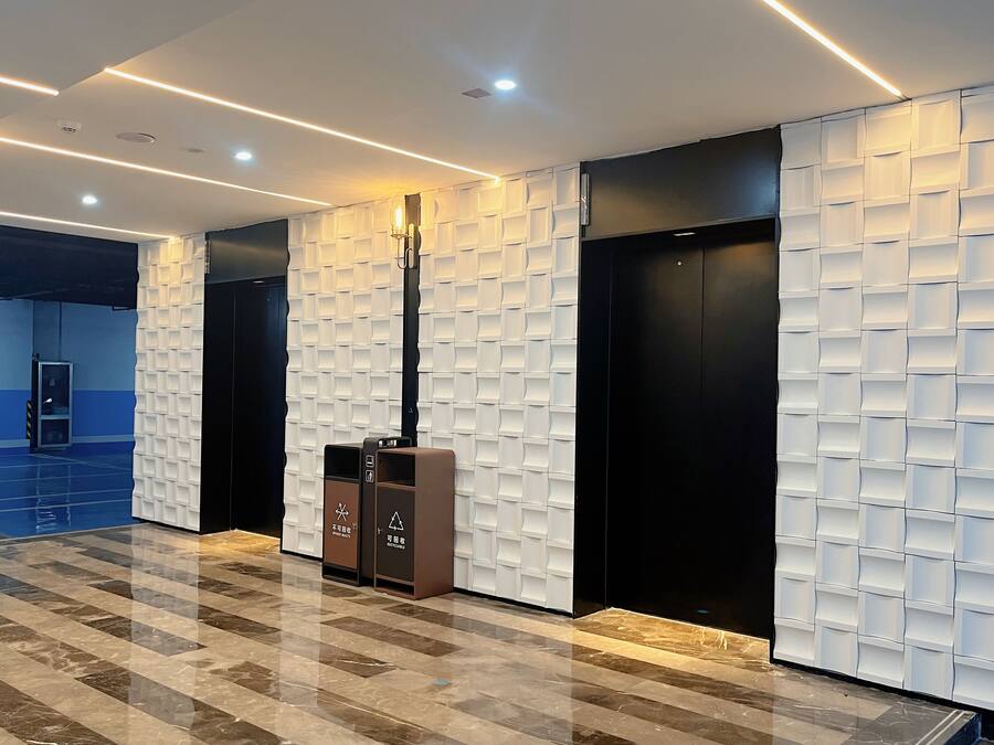 A modern lobby with a marble floor, a tall column with a light fixture, and two elevators with a unique textured wall design.
