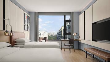 A modern hotel room with two beds, a desk, a chair, a bench, and a large TV.