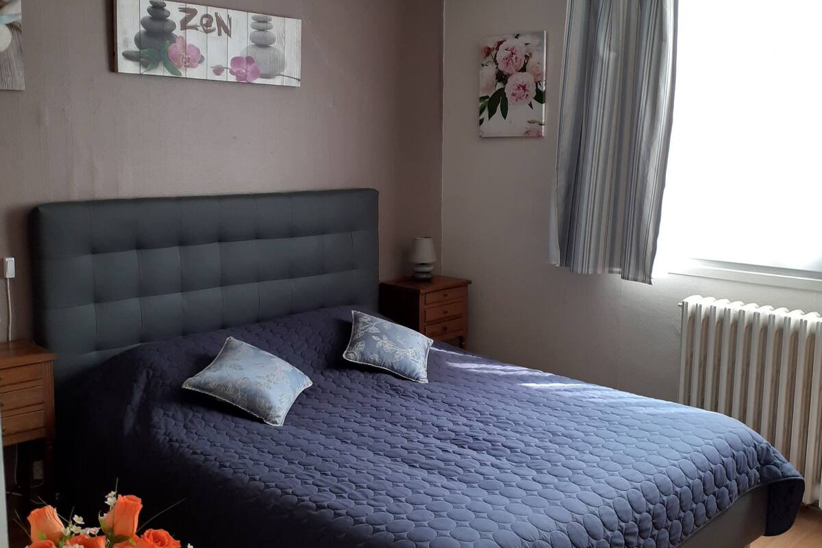 Quarto com uma cama grande, uma mesa de cabeceira, um criado-mudo, um vaso de flores e uma janela com cortinas.