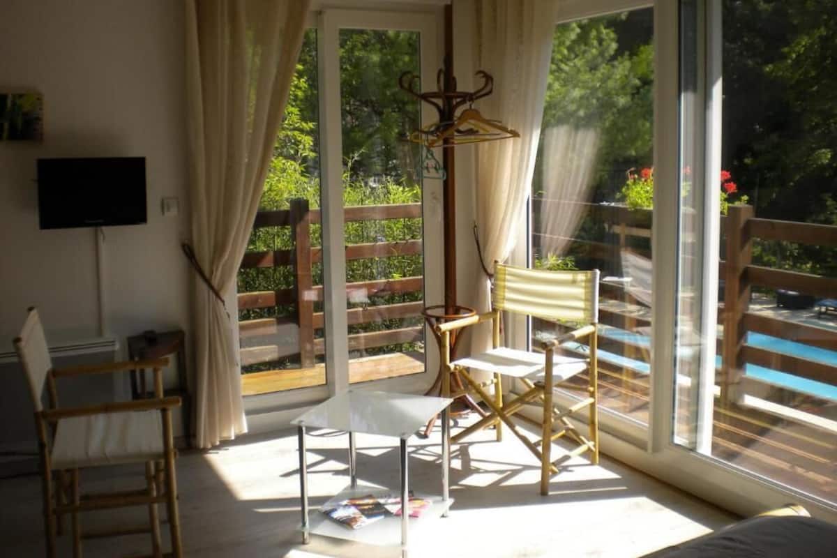 Um quarto com uma grande porta de vidro deslizante que dá para uma varanda, uma mesa com cadeiras, um cabideiro e uma televisão na parede.