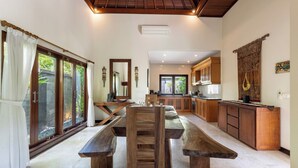 A spacious kitchen with wooden cabinets, a large dining table, and a view of the outdoors through sliding glass doors. - Senang Villa by Betterplace (Canggu)