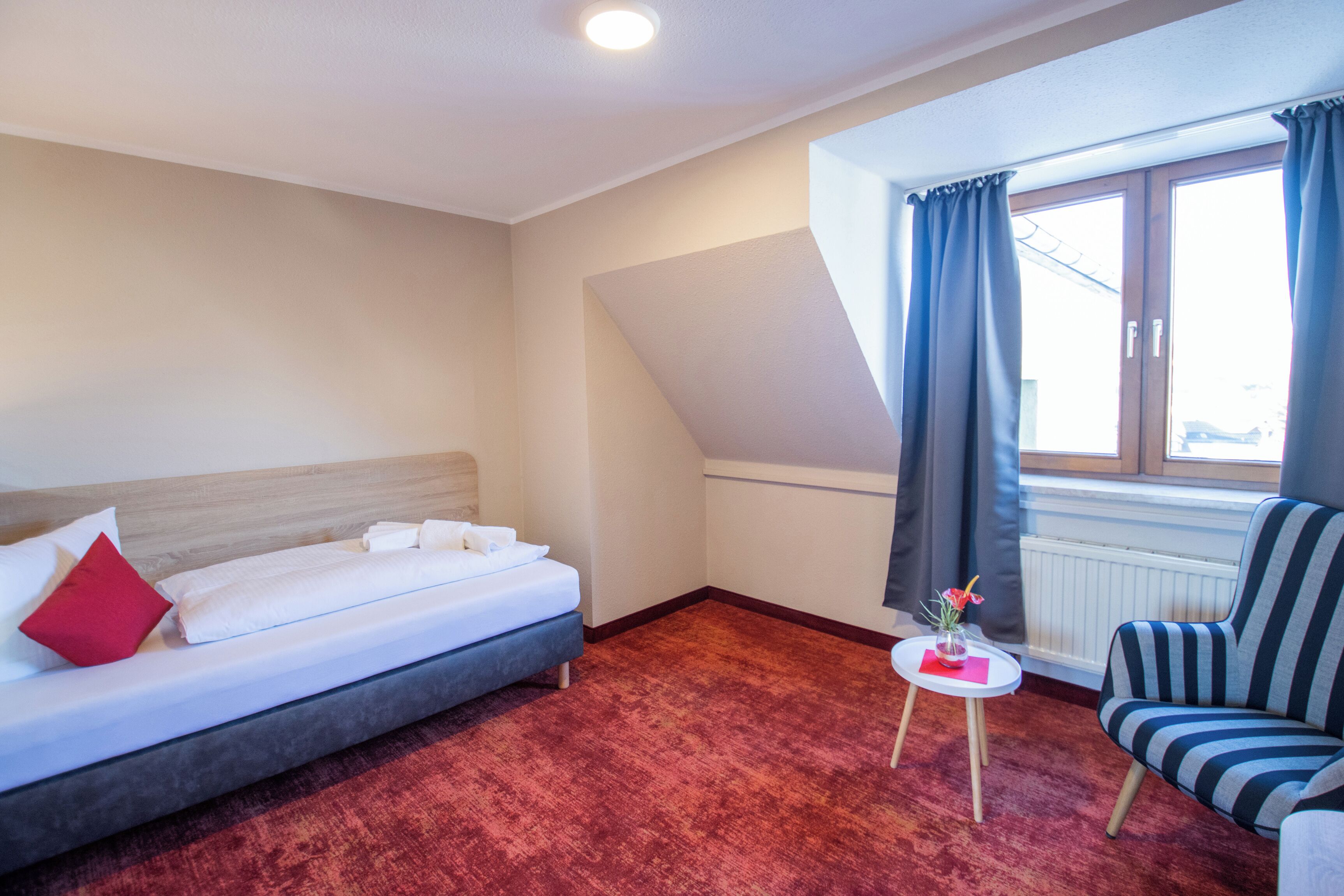 Quarto de hotel com uma cama, uma mesinha com um vaso, uma cadeira e uma janela com cortinas.