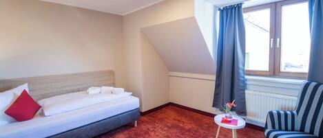 A hotel room with a bed, a small table with a vase, a chair, and a window with curtains.