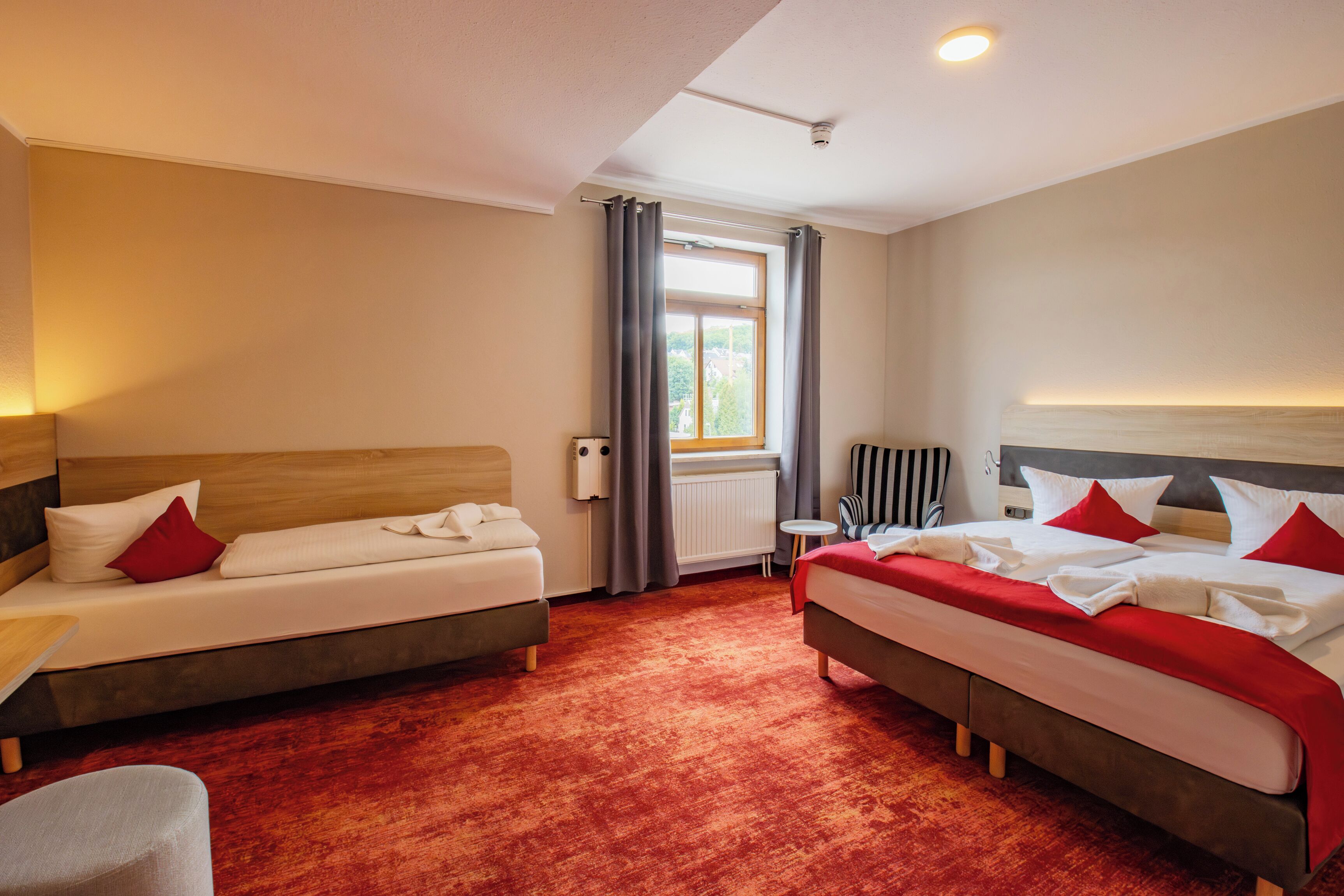 A hotel room with two beds, a small table, a radiator, and a window with curtains.