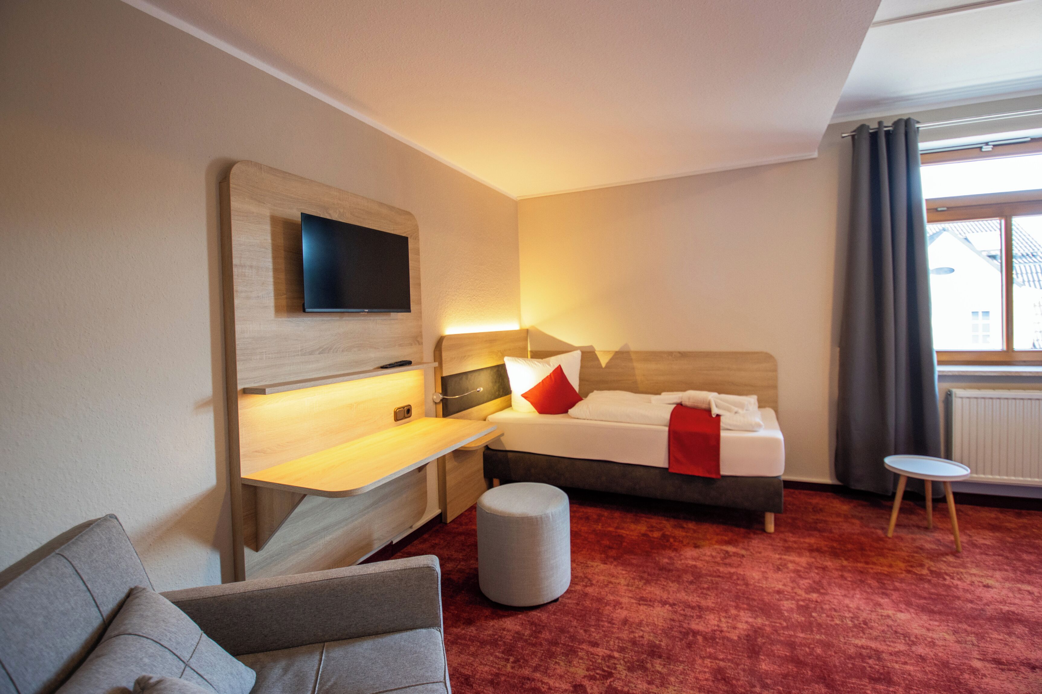Habitación de hotel con cama, sofá, una mesita, televisor montado en un panel de madera y ventana con cortinas.