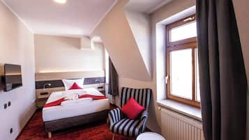 A hotel room with a bed, a chair, a small table, a window with curtains, and a radiator.
