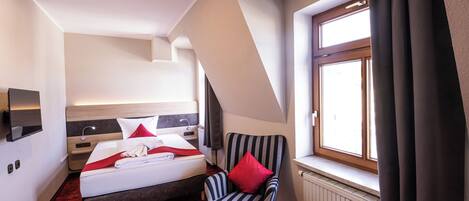 Habitación de hotel con cama, una silla, una mesita, ventana con cortinas y un radiador.
