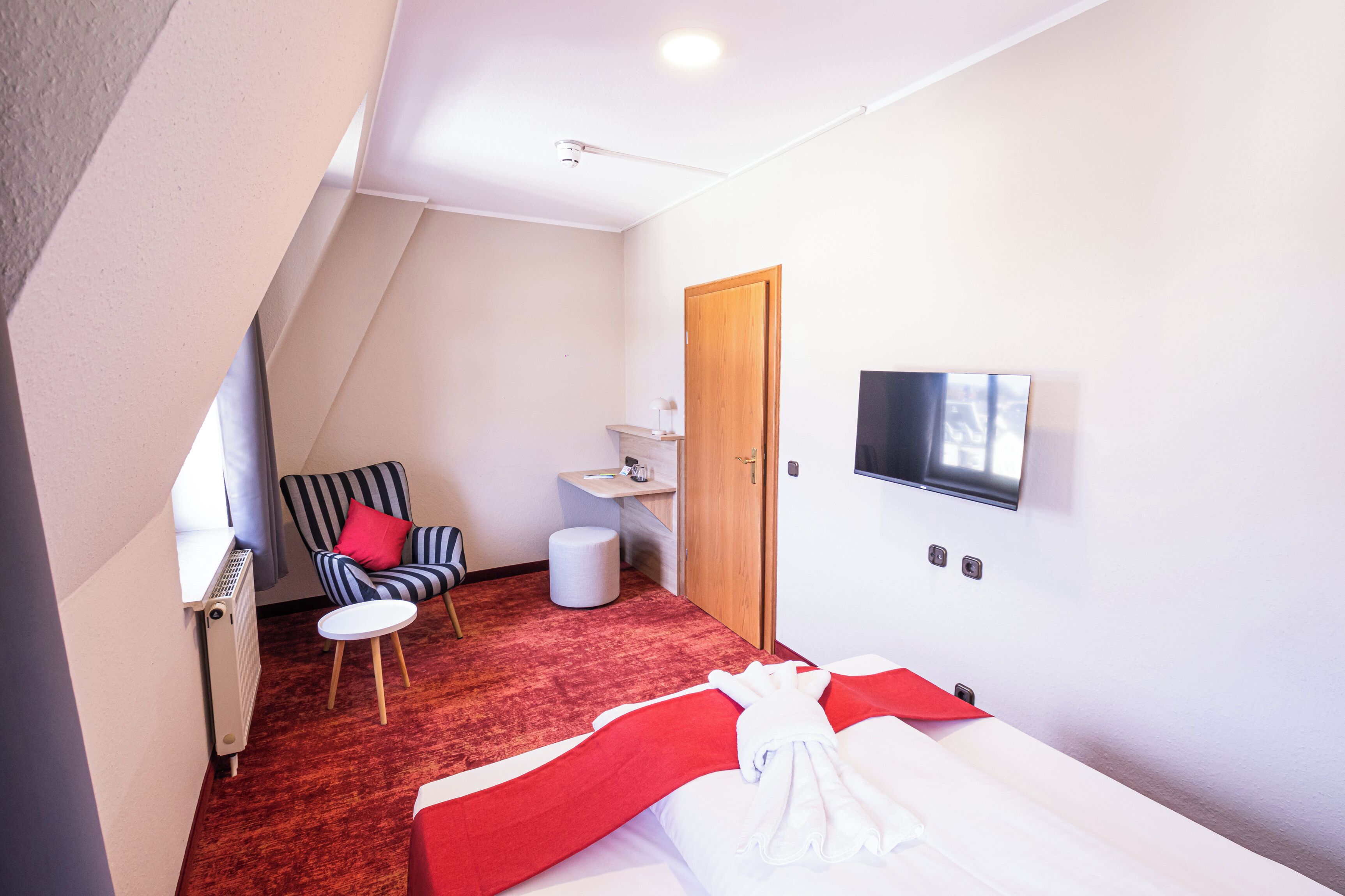 A hotel room with a bed, a TV mounted on the wall, a small table, a chair, and a radiator.
