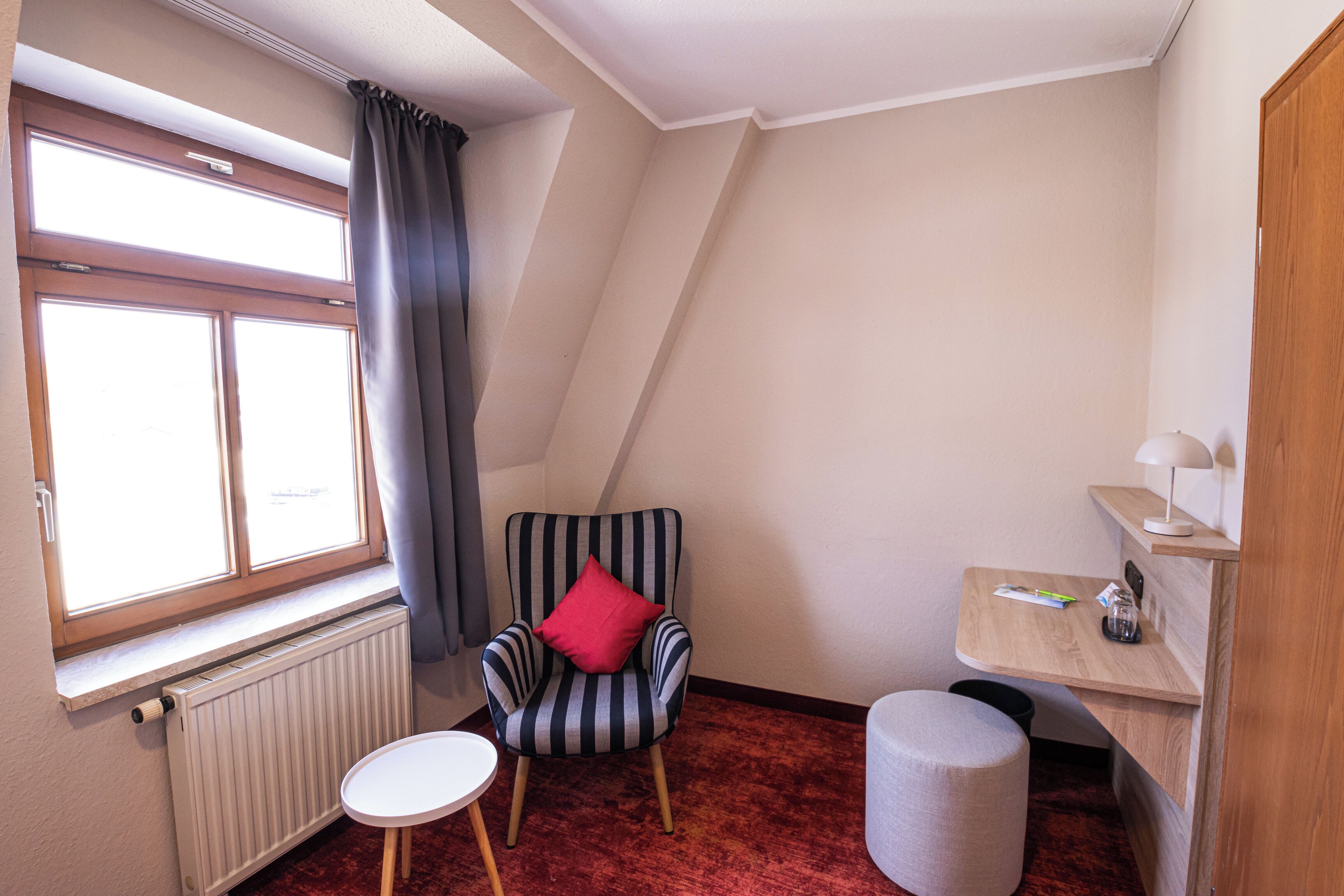Um quarto pequeno e acolhedor com uma poltrona listrada, uma mesa redonda pequena, um radiador e uma janela com cortinas.