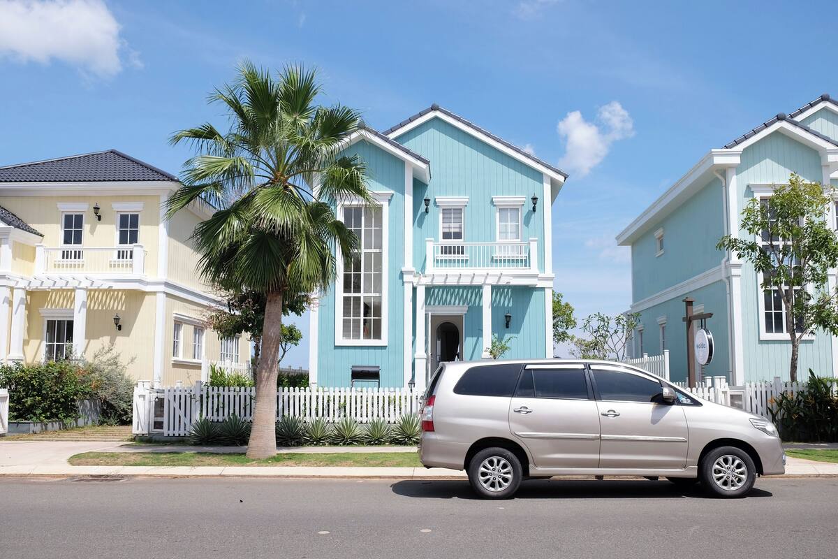 Ein beiges Minivan steht auf einer Straße vor einem hellblauen Haus mit weißem Zaun.