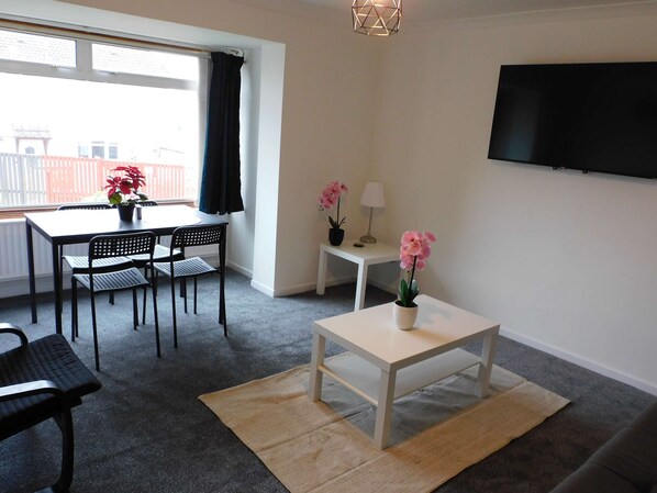 A modern living room with a dining area, a flat-screen TV mounted on the wall, and a window with curtains. - Newly refurbished 3 bedroom home in East Kilbride - 52 (East Kilbride)