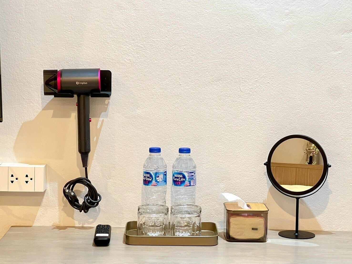 Habitación de hotel con secador de pelo, dos botellas de agua, un control remoto, una bandeja con vasos, una caja de pañuelos de papel y un espejo redondo.