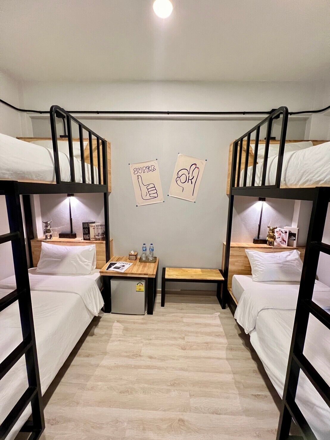 A dormitory room with bunk beds, a small desk with a chair, a nightstand, and two wall decorations.