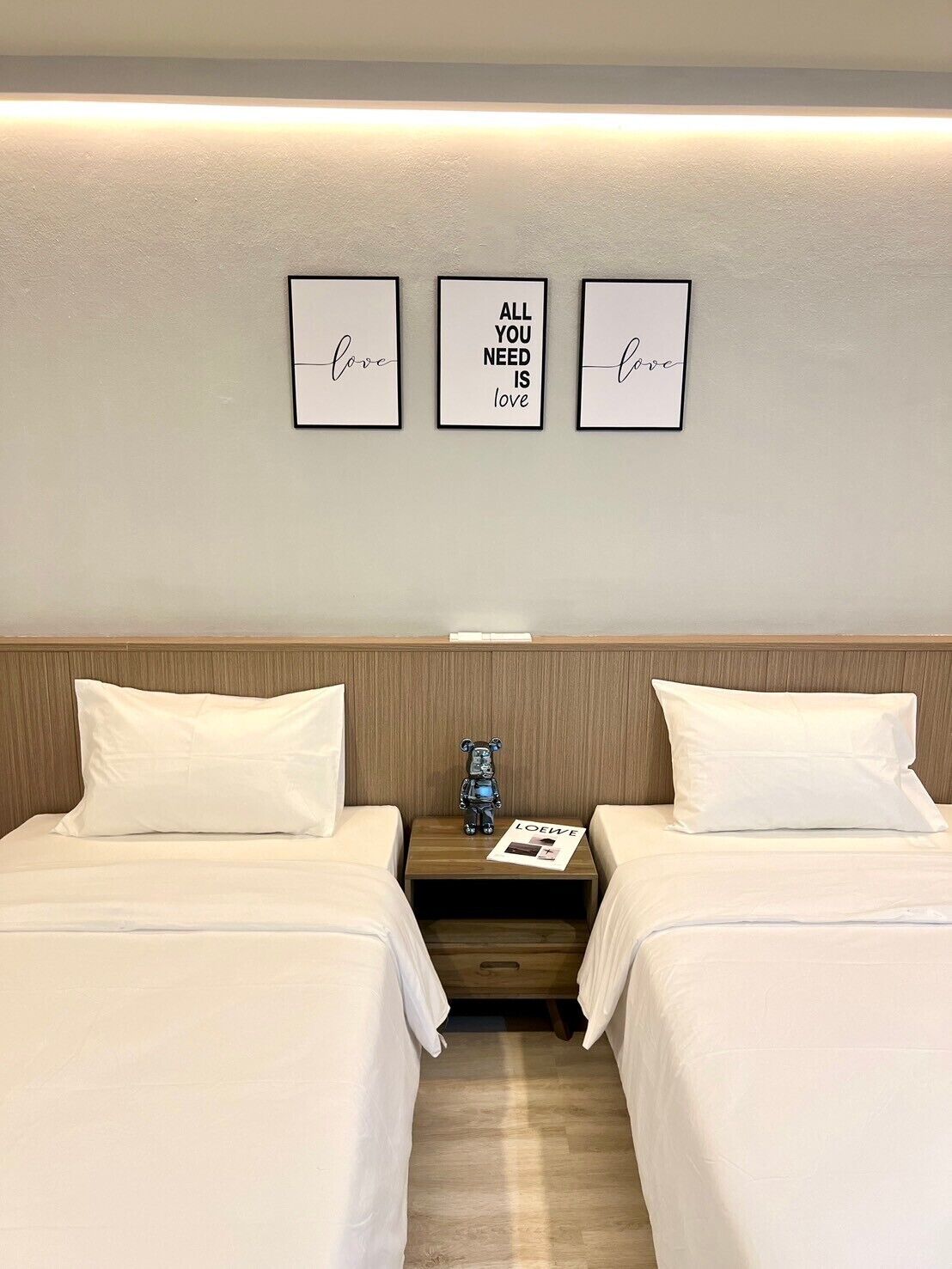 Une chambre d’hôtel avec deux lits, une table de chevet sur laquelle sont posés un ours en peluche et un magazine, et trois citations inspirantes encadrées accrochées au mur.