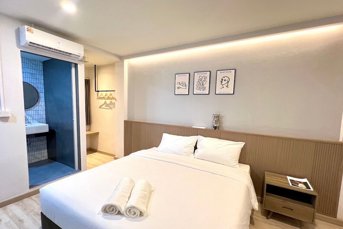A hotel room with a bed, a nightstand, a wall-mounted air conditioner, a bathroom with a blue-tiled shower, and three framed artworks on the wall.