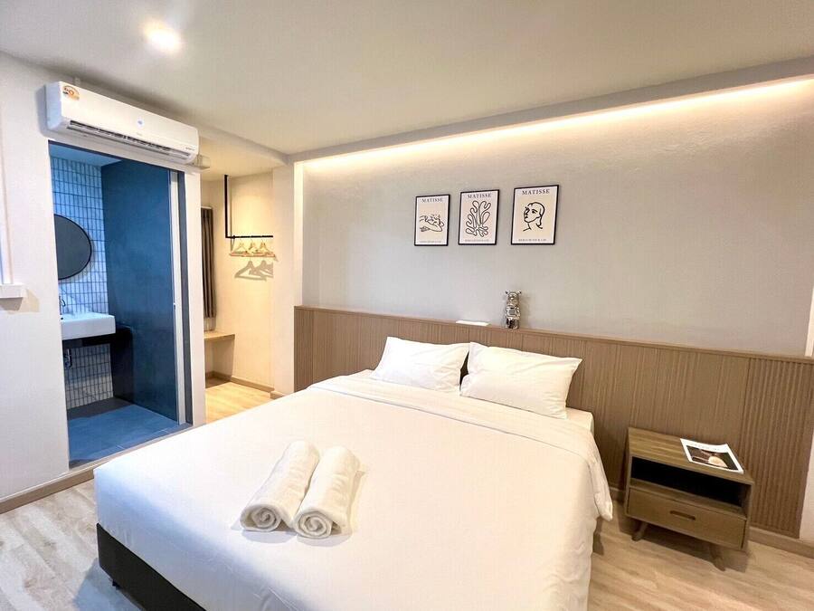 A hotel room with a bed, a nightstand, a wall-mounted air conditioner, a bathroom with a blue-tiled shower, and three framed artworks on the wall.