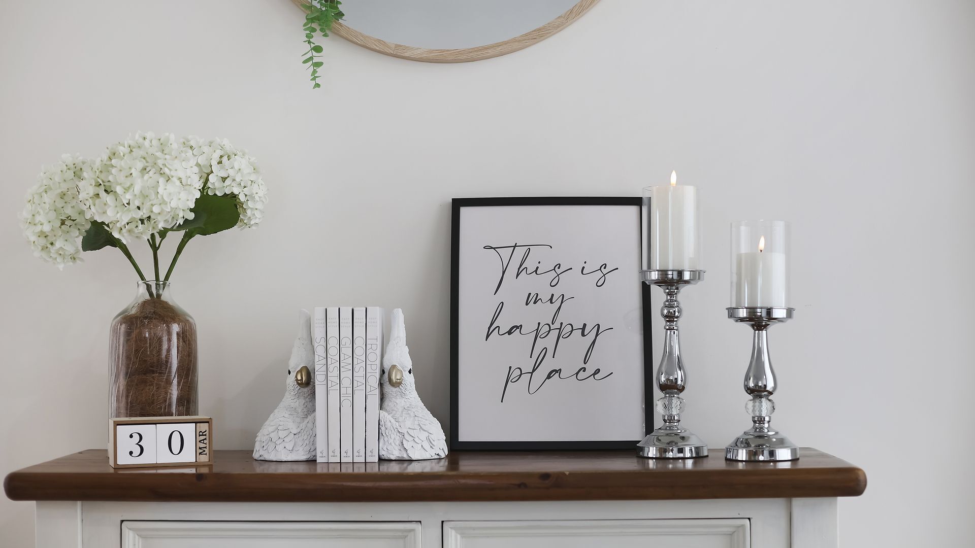 A wooden mantel with a framed sign, a vase of white flowers, a decorative white object, and two lit candles.