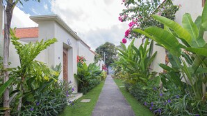 Un sendero bordeado de exuberante vegetación y plantas florecientes, que conduce a un edificio con fachada blanca y techo de tejas rojas.