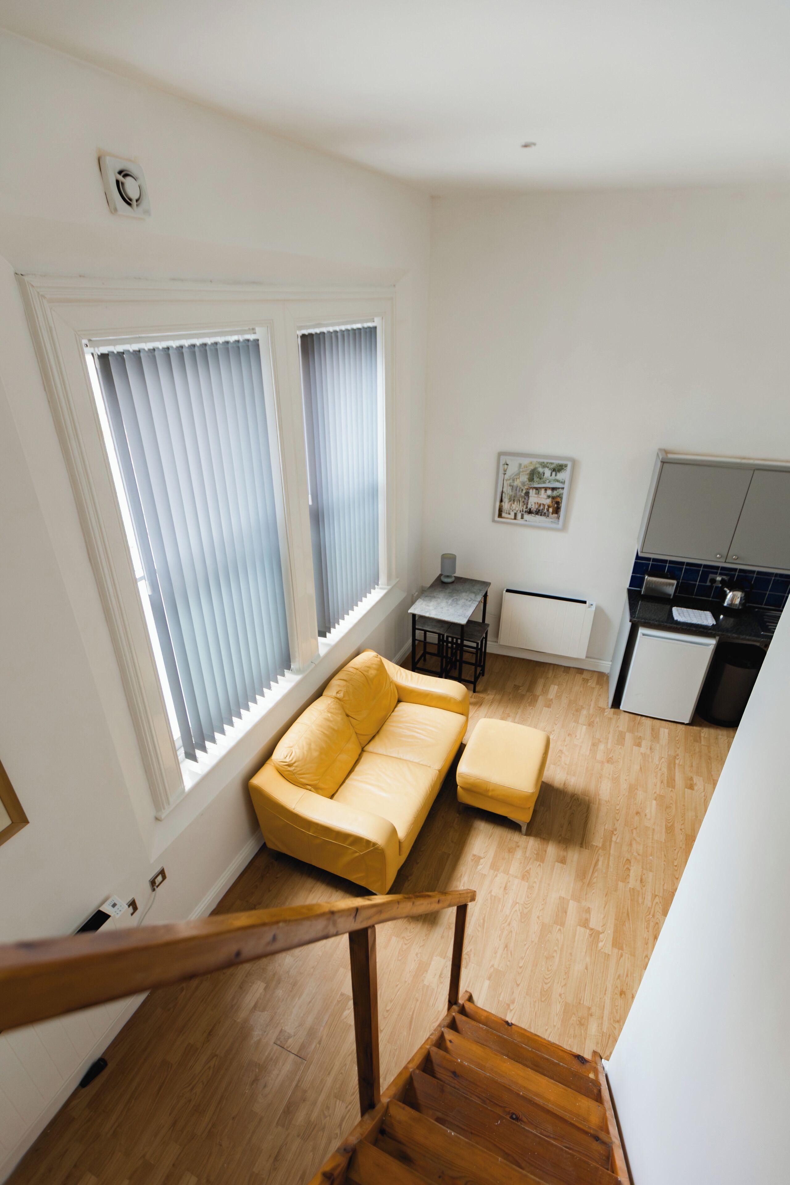 A modern apartment with a wooden staircase, a yellow sofa, a small kitchen, and large windows with blinds.