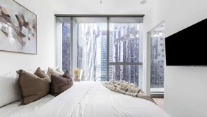 Ein modernes Schlafzimmer mit einem großen Bett, Blick auf die Stadt durch raumhohe Fenster und einem an der Wand montierten Fernseher.