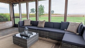 Ein überdachter Außenbereich mit einer Couch und einem Couchtisch, große Fenster mit Blick auf ein Feld und Bäume.