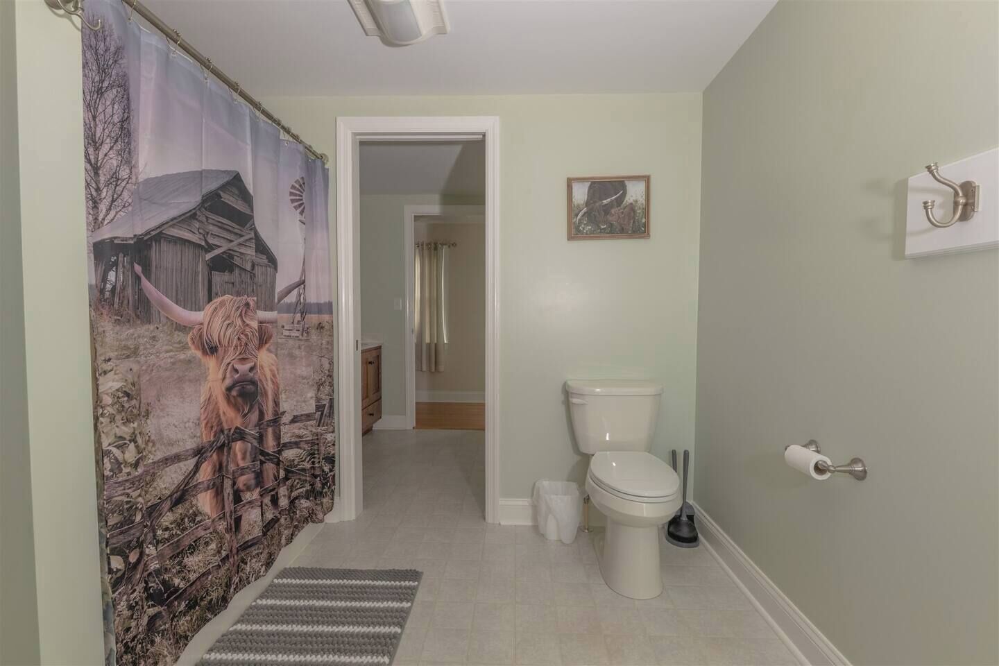 Une salle de bain avec des toilettes, un lavabo, un rideau de douche orné d’une vache et un petit tableau accroché au mur.