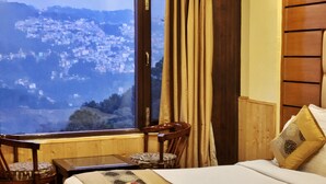 A hotel room with a bed, a table with a plate of fruit, a chair, and a view of a cityscape through a window. - Hotel Snow Lotus (Shimla)