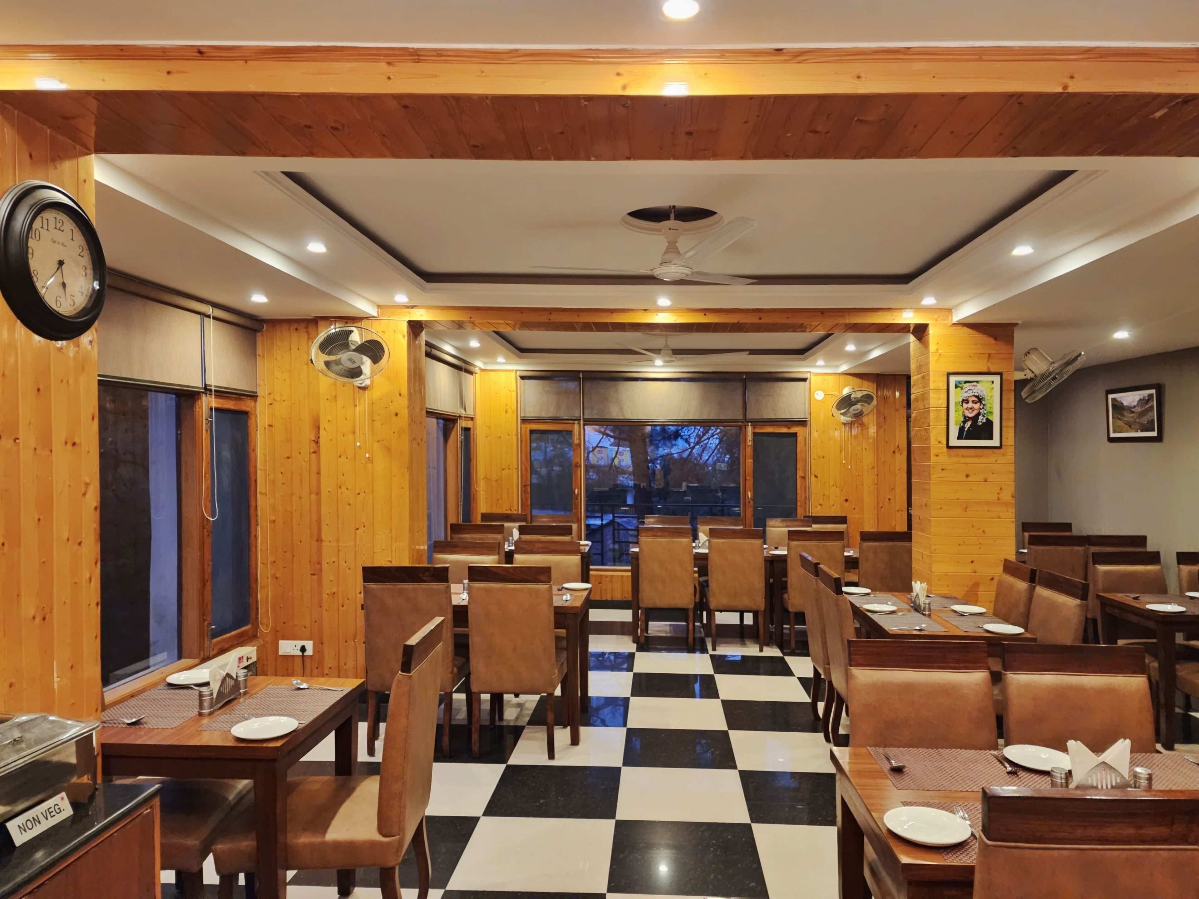 A restaurant with wooden walls and a checkered floor, featuring tables and chairs, a clock, and ceiling fans.