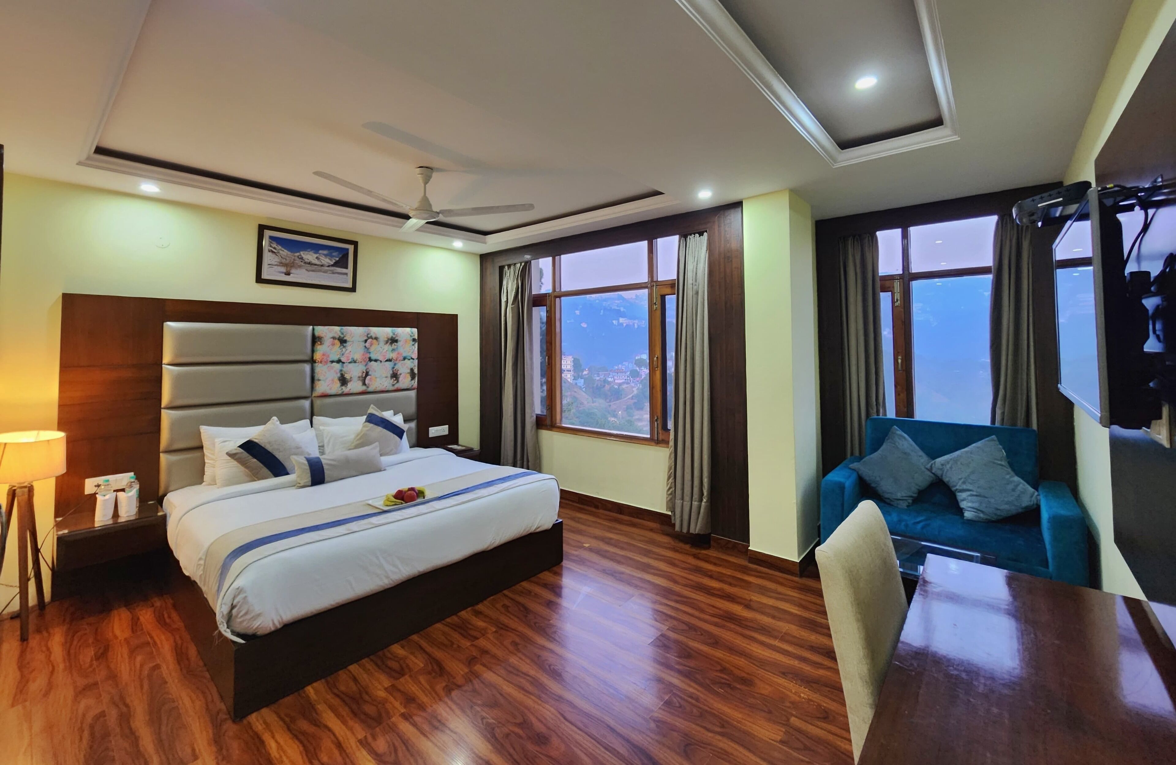 A hotel room with a large bed, a blue sofa, a dining table, and a view of the city through large windows.