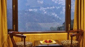 A room with a view of a hillside town through a window, wooden furniture, and a plate of fruit on a table. - Hotel Snow Lotus (Shimla)