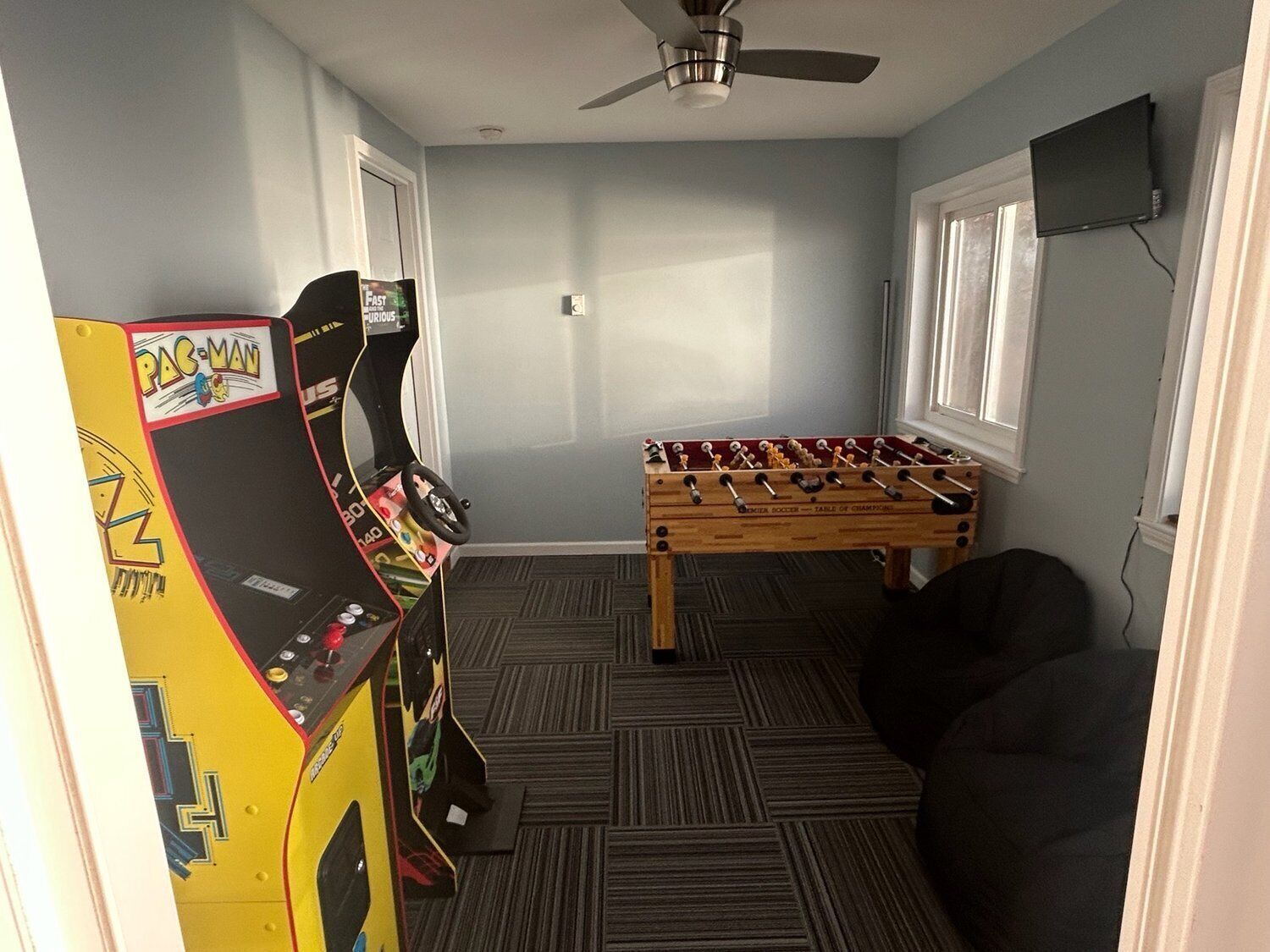 A room with a foosball table, two arcade machines, a ceiling fan, and a TV mounted on the wall.