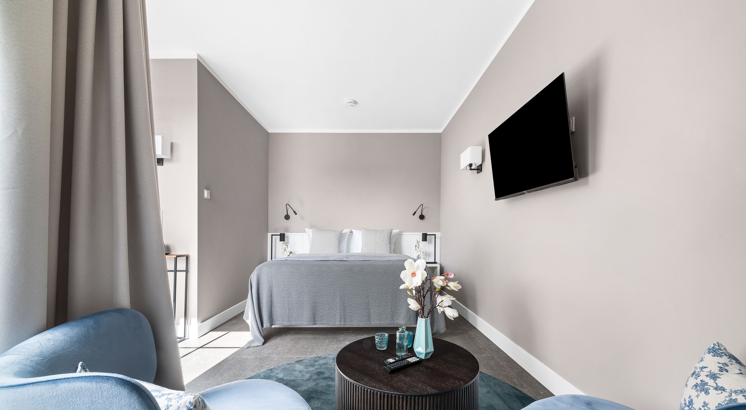 Une chambre d’hôtel moderne avec un lit, un téléviseur fixé au mur, une table basse ronde et un fauteuil bleu.