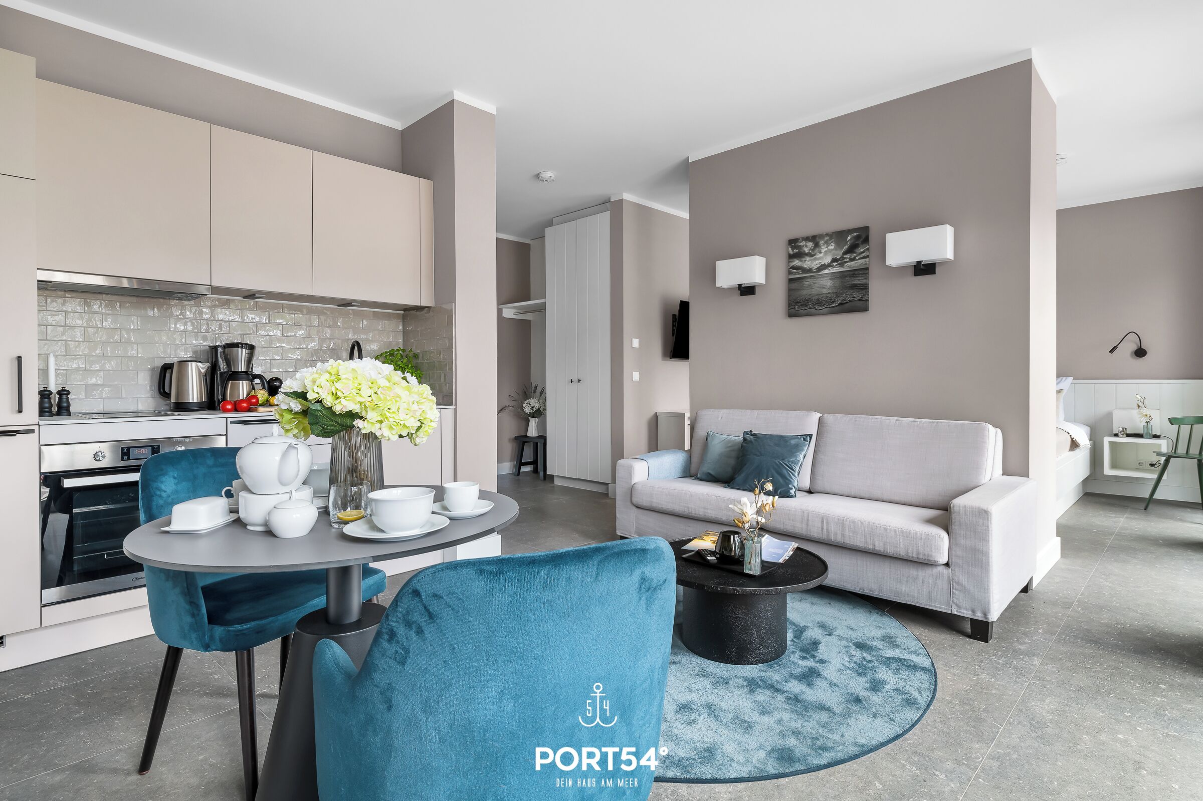 Ein modernes Wohnzimmer mit grauem Sofa, einem Couchtisch und einem Essbereich mit Tisch und Stühlen. Im Hintergrund befindet sich eine Küche mit Schränken und einem Herd.