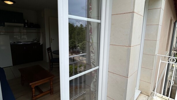 A room with a dining table, a kitchen area, and a balcony with a view of greenery. - T2 Apartment with Balcony and Shared Pool, Near Spa (La Roche-Posay)