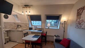 A compact kitchen with a dining table and chairs, a sofa with red cushions, and a large window with a view of buildings.