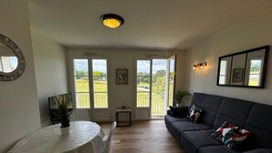 A spacious living area with a dark-colored sofa, a round table with a white tablecloth, and a view through large glass doors to an outdoor garden. - T2 Apartment with Wi-Fi and Pool 200m from Spas (La Roche-Posay)