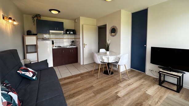 A modern living room with a dark sofa, a dining table with chairs, a kitchen area, and a TV on a stand. - T2 Apartment with Wi-Fi and Pool 200m from Spas (La Roche-Posay)
