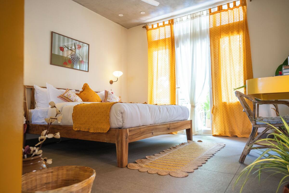 A bedroom with a wooden bed, a bedside table, a chair, a lamp, and a window with curtains.