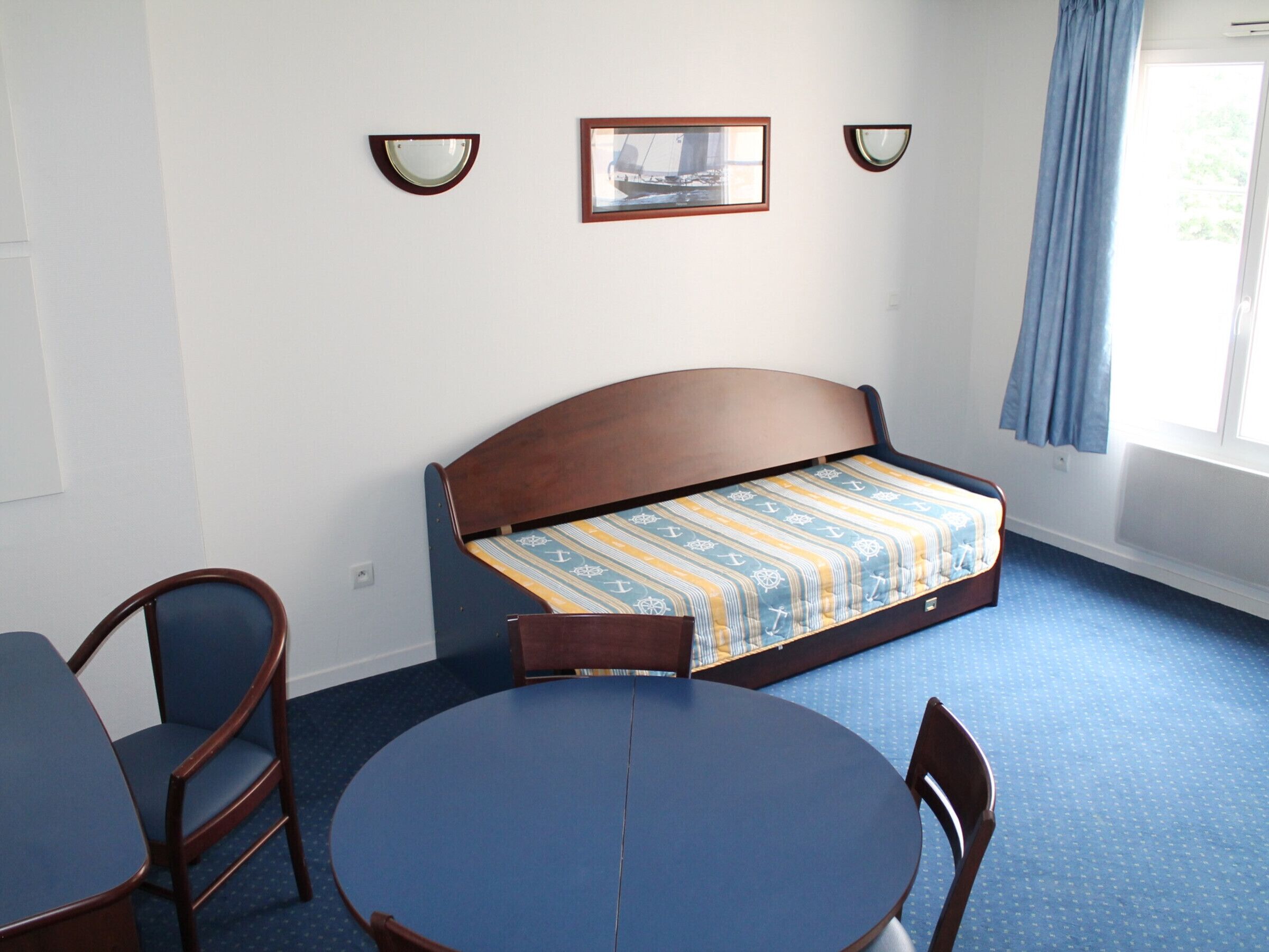 A room with a bed, a round table, two chairs, a window with a curtain, and wall-mounted lights.