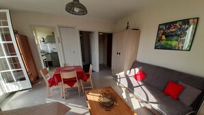 Ein Wohnzimmer mit einer Couch, einem Holztisch und einem Esstisch mit Stühlen. Im Hintergrund ist eine Küche zu sehen.
