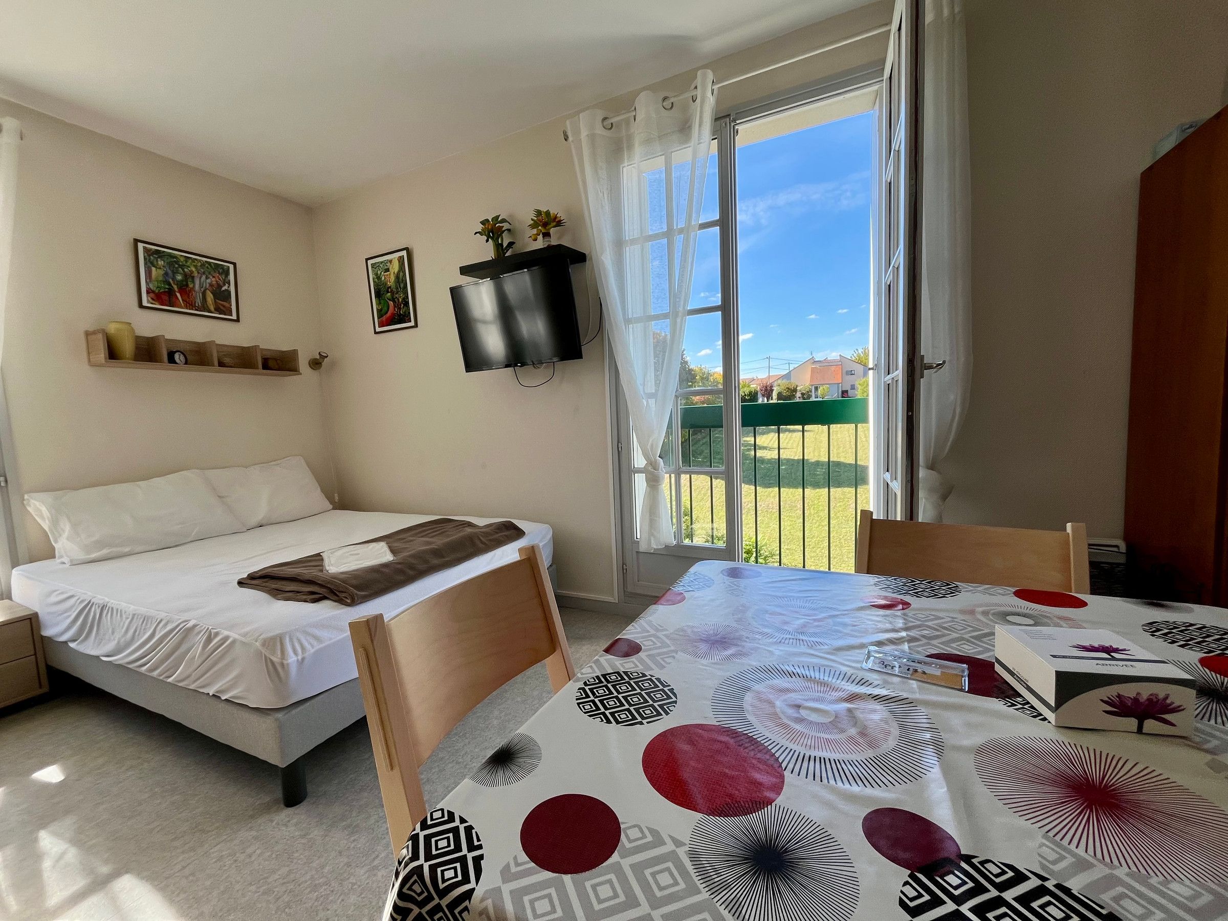 A bedroom with a bed, a table with a patterned tablecloth, a TV mounted on the wall, and a view of the outside through a window.