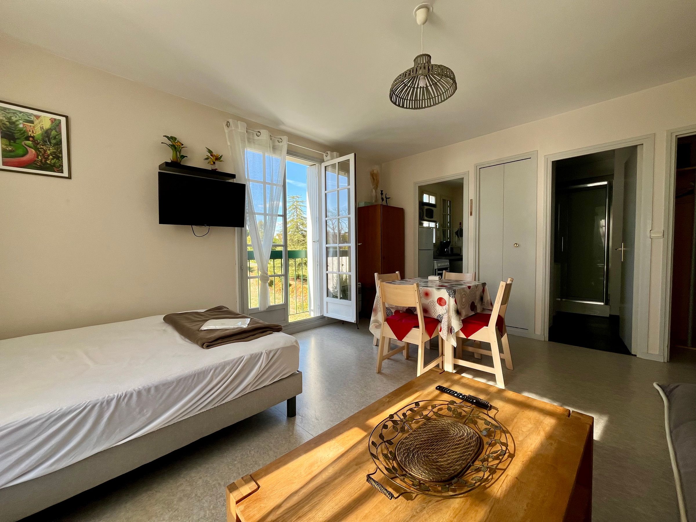 A room with a bed, a dining table with chairs, a TV mounted on the wall, and a view through a window.