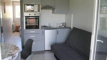 Een compacte kitchenette met een magnetron, oven en spoelbak. Er is een bank en een eettafel met stoelen.