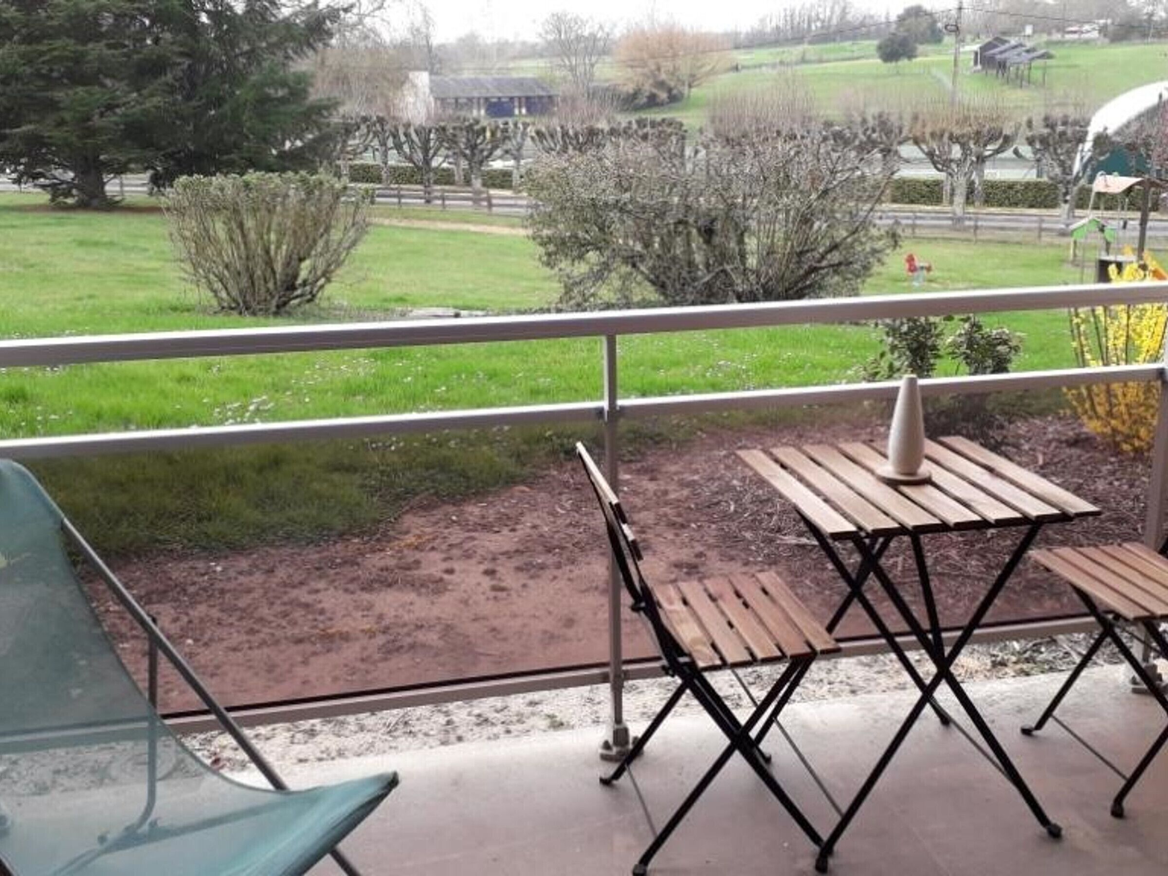 A balcony with a wooden table and chairs, a metal railing, and a view of a green field and trees.