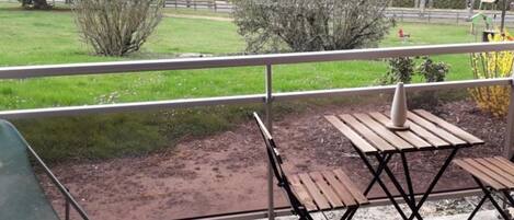 A balcony with a wooden table and chairs, a metal railing, and a view of a green field and trees.