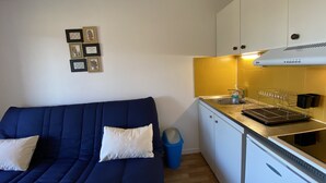 A compact living space with a sofa, a small kitchenette, and a wall-mounted shelf with framed pictures. - 1-bedroom near Connétable Thermal Baths with balcony and Wi-Fi (La Roche-Posay)