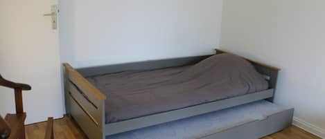 A small, single-bed room with a wooden floor, a simple bed frame, and a door with a handle.