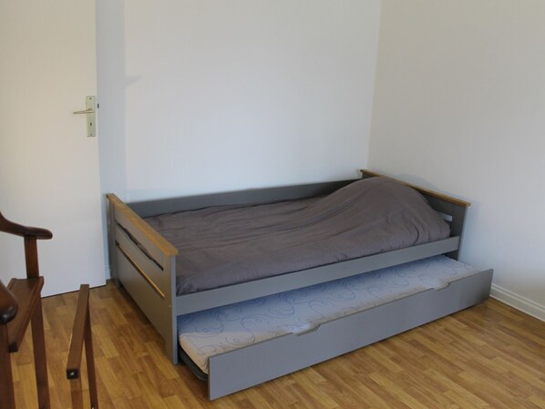 Una habitación pequeña con cama individual, piso de madera, sencillo cabecero y una puerta con picaporte.